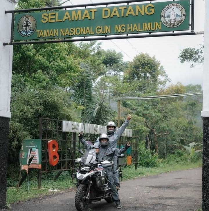 Dua pengendara motor mengenakan jaket dan helm berpose di bawah gerbang masuk Taman Nasional Gunung Halimun Salak yang dikelilingi pepohonan hijau.