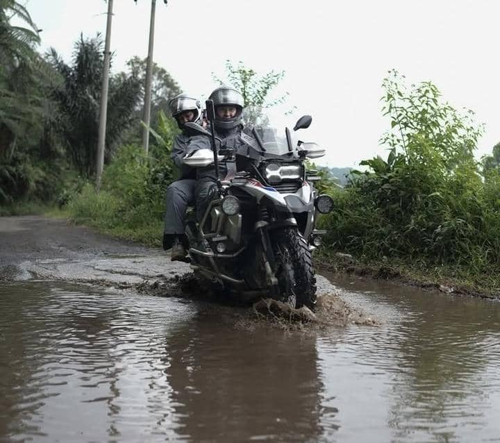 Dua pengendara motor trail melintasi jalan berlumpur dan berair di kawasan hijau dengan mengenakan jaket dan helm pelindung.