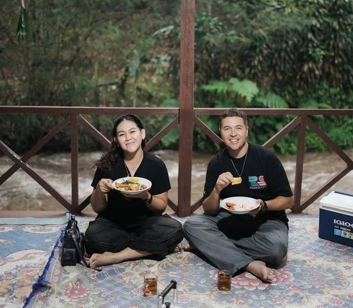 Dua orang duduk bersila di teras kayu dekat sungai sambil menikmati makanan di piring, dengan latar pepohonan hijau yang rimbun.
