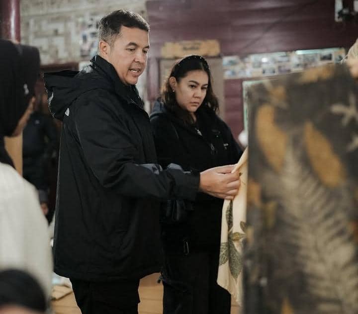 Seorang pria dan wanita mengenakan jaket hitam sedang memperhatikan kain bermotif daun di dalam ruangan dengan pencahayaan hangat.