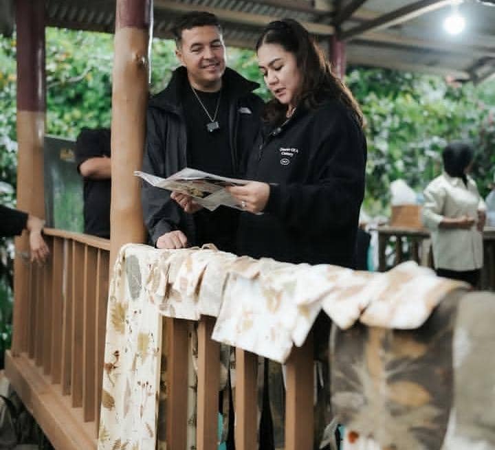 Seorang pria dan wanita mengenakan jaket hitam berdiri di teras kayu sambil melihat lembaran kertas, dengan kain bermotif tergantung di pagar.