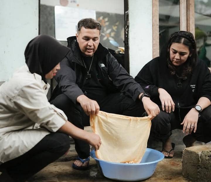 Tiga orang duduk bersama di depan bangunan sambil memegang kain berwarna krem di atas baskom biru berisi air.