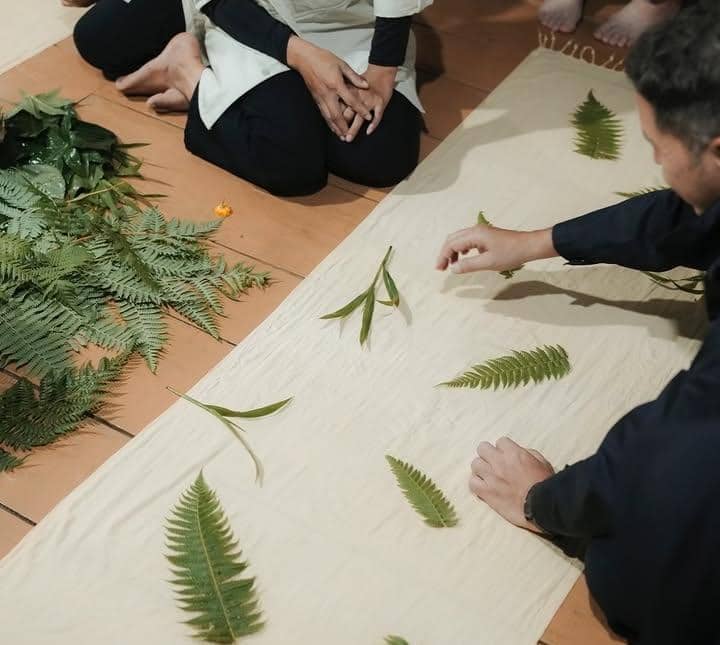 Beberapa orang duduk di lantai sambil menyusun daun pakis dan dedaunan hijau di atas kain putih panjang untuk kegiatan kreatif.