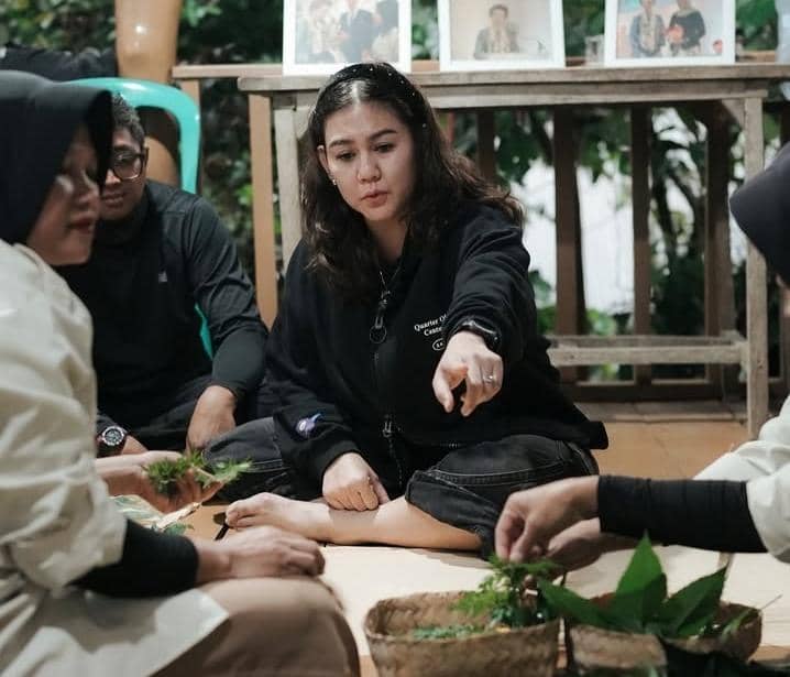 Seorang perempuan berjaket hitam duduk bersama beberapa orang di lantai, menunjuk ke arah daun dan bahan alami di depan mereka.