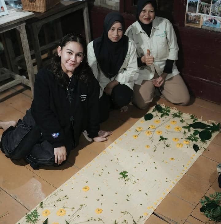 Tiga perempuan duduk di lantai di samping kain panjang berhiaskan bunga kuning dan daun hijau di dalam ruangan kayu.