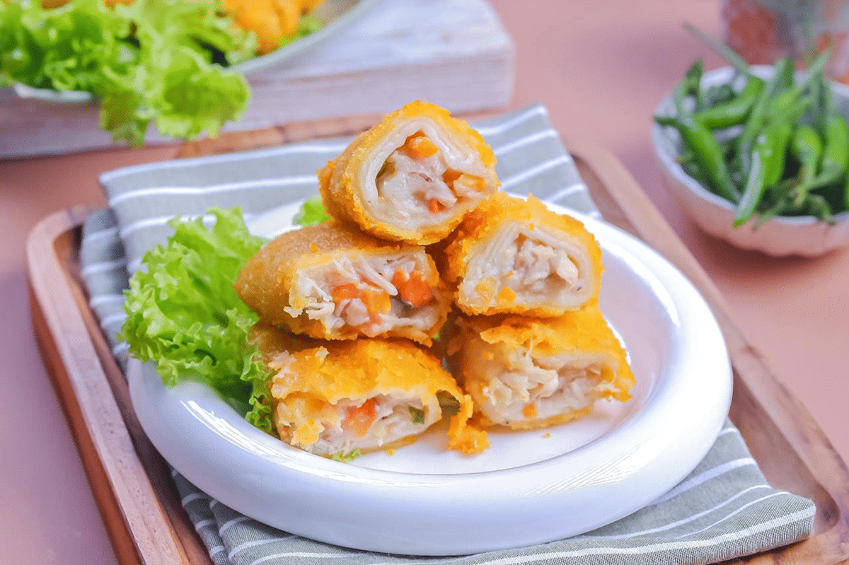 Risoles isi ragout dengan kulit renyah berwarna keemasan disajikan di atas piring putih bersama daun selada segar.