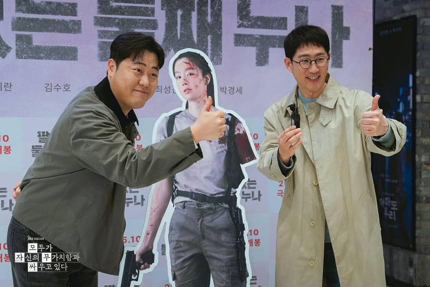 Dua pria berpose dengan jempol di depan poster drama We Are All Trying Here yang menampilkan karakter wanita memegang pistol.
