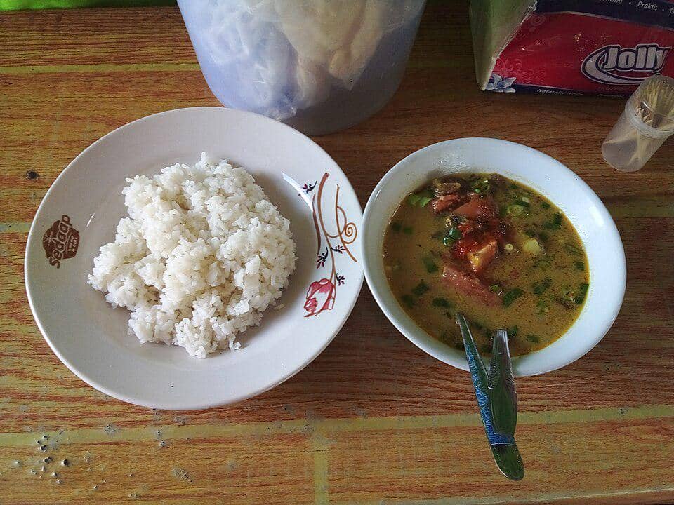Semangkuk soto tangkar daging kambing berkuah kuning dengan potongan daging dan sambal, disajikan bersama sepiring nasi putih.