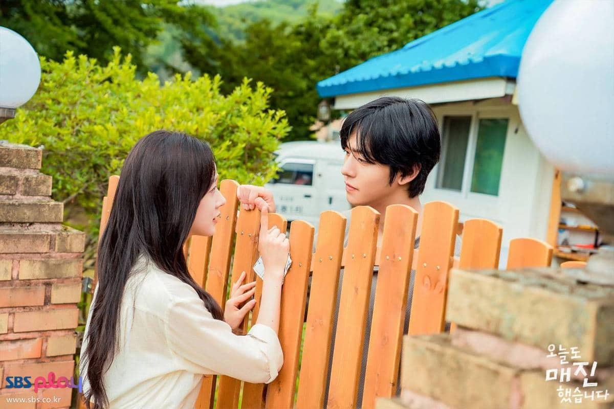 Dua karakter drama Sold Out On You berbincang di pagar kayu dengan latar rumah dan pepohonan hijau di siang hari.