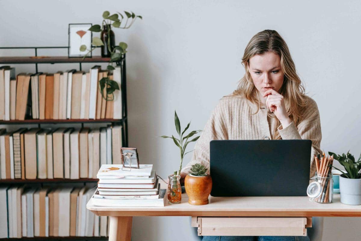 Seorang perempuan duduk di meja kerja dengan laptop di depannya, dikelilingi buku, tanaman hias, dan alat tulis di ruang kerja minimalis.