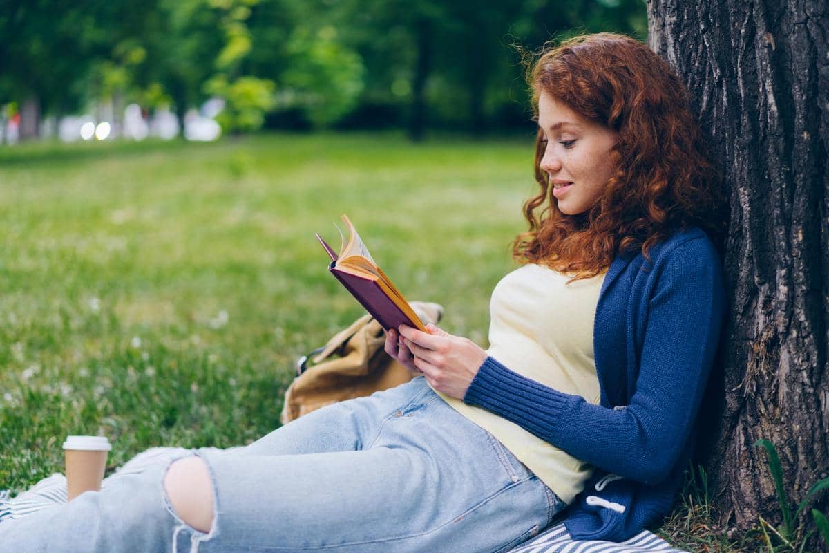 Seorang perempuan duduk bersandar di bawah pohon sambil membaca buku di taman hijau dengan suasana tenang dan santai.