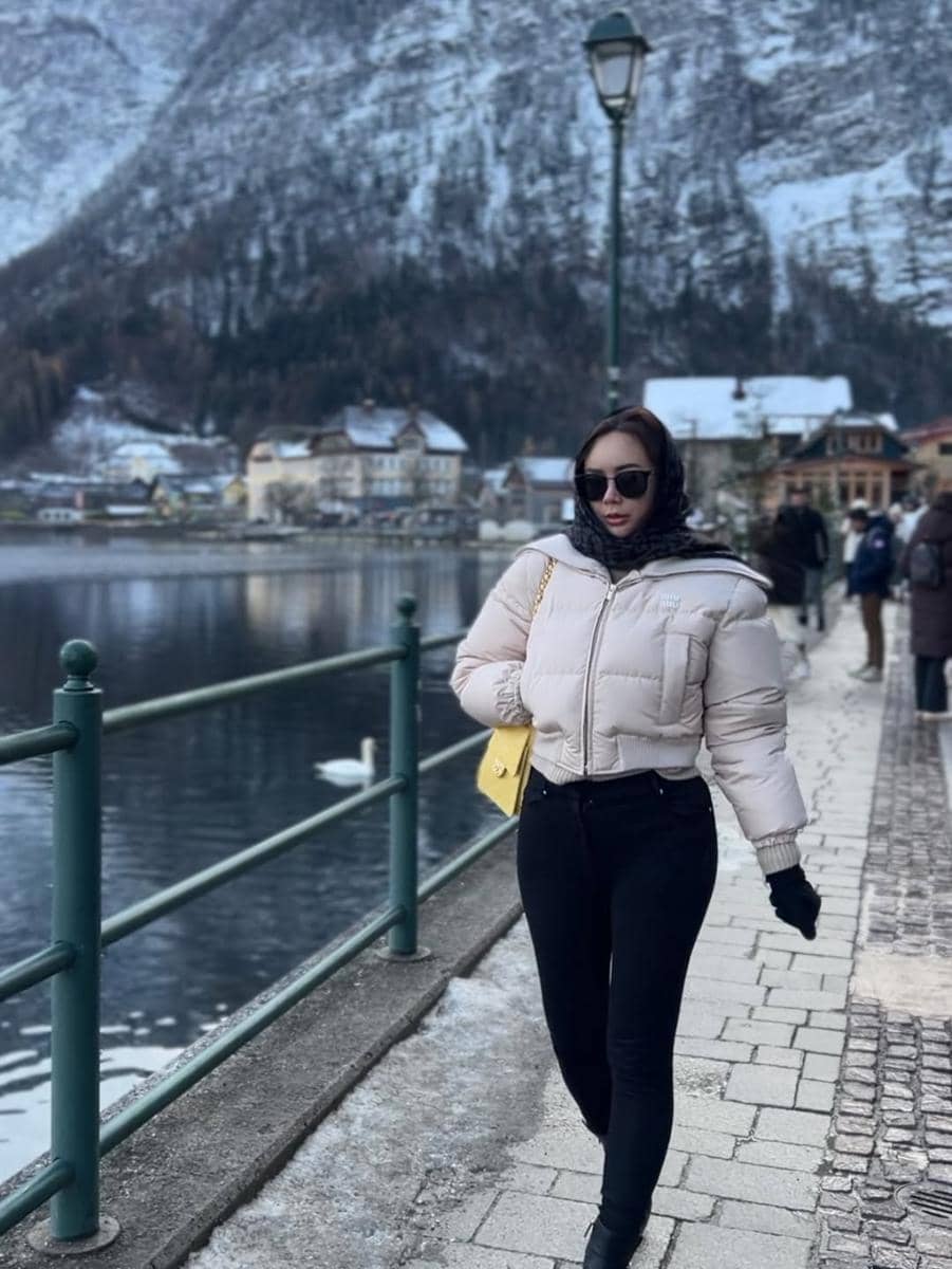 Seorang wanita mengenakan jaket tebal dan kacamata hitam berjalan di tepi danau dengan latar pegunungan bersalju di Austria.