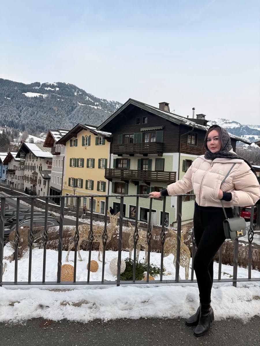 Seorang wanita mengenakan jaket tebal dan syal berdiri di depan pagar dengan latar bangunan bergaya Eropa dan pegunungan bersalju di Austria.