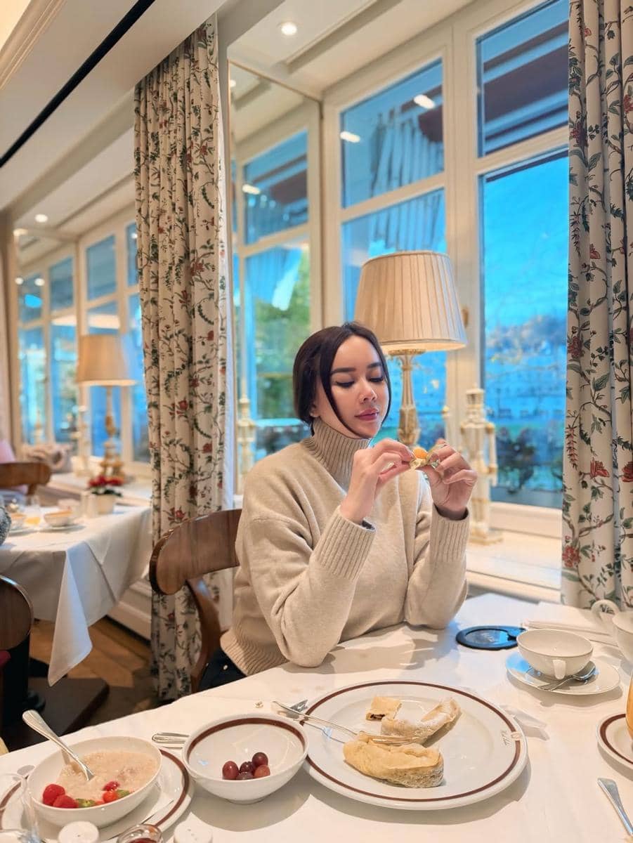 Seorang perempuan mengenakan sweater krem duduk di restoran elegan dengan jendela besar, menikmati hidangan sarapan di Austria.