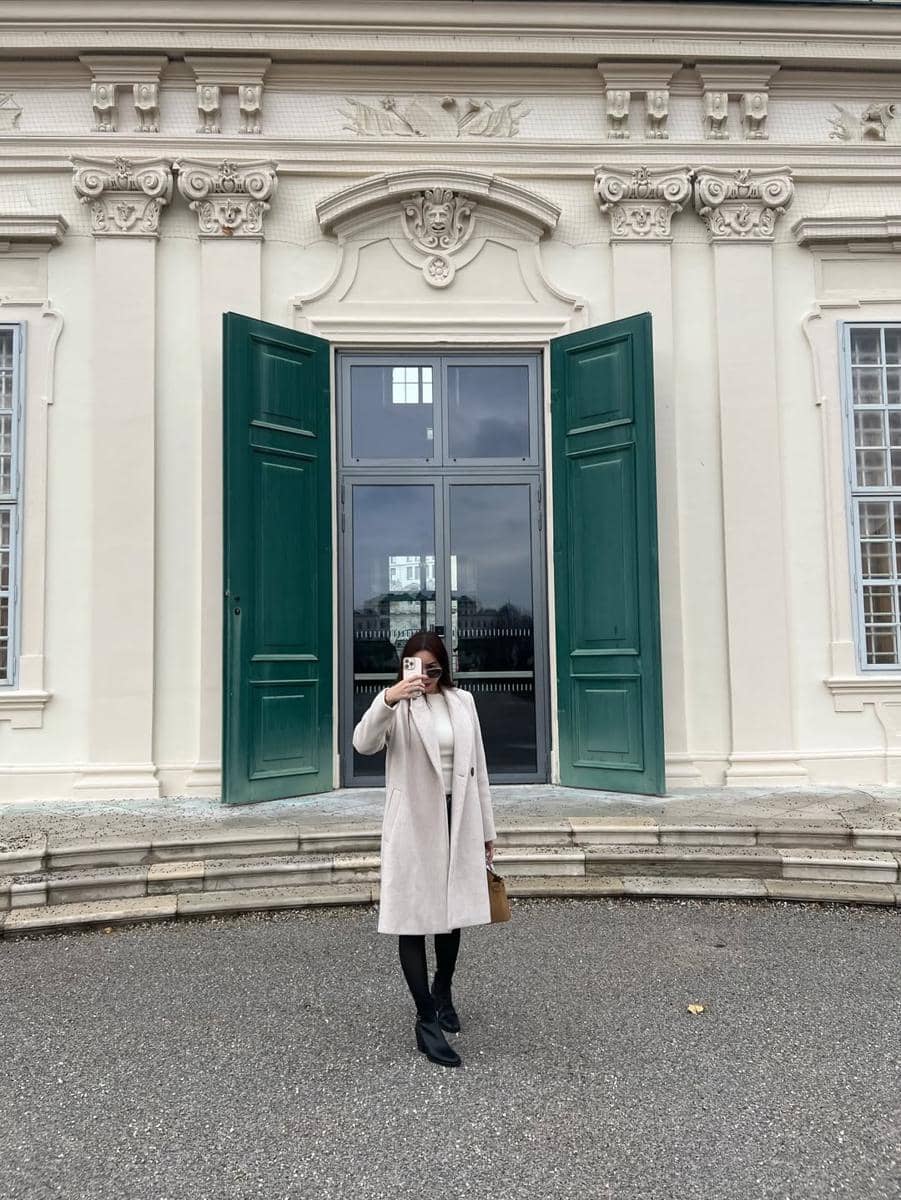 Seorang wanita berpose sambil memegang ponsel di depan bangunan bergaya klasik dengan pintu hijau besar di Austria.