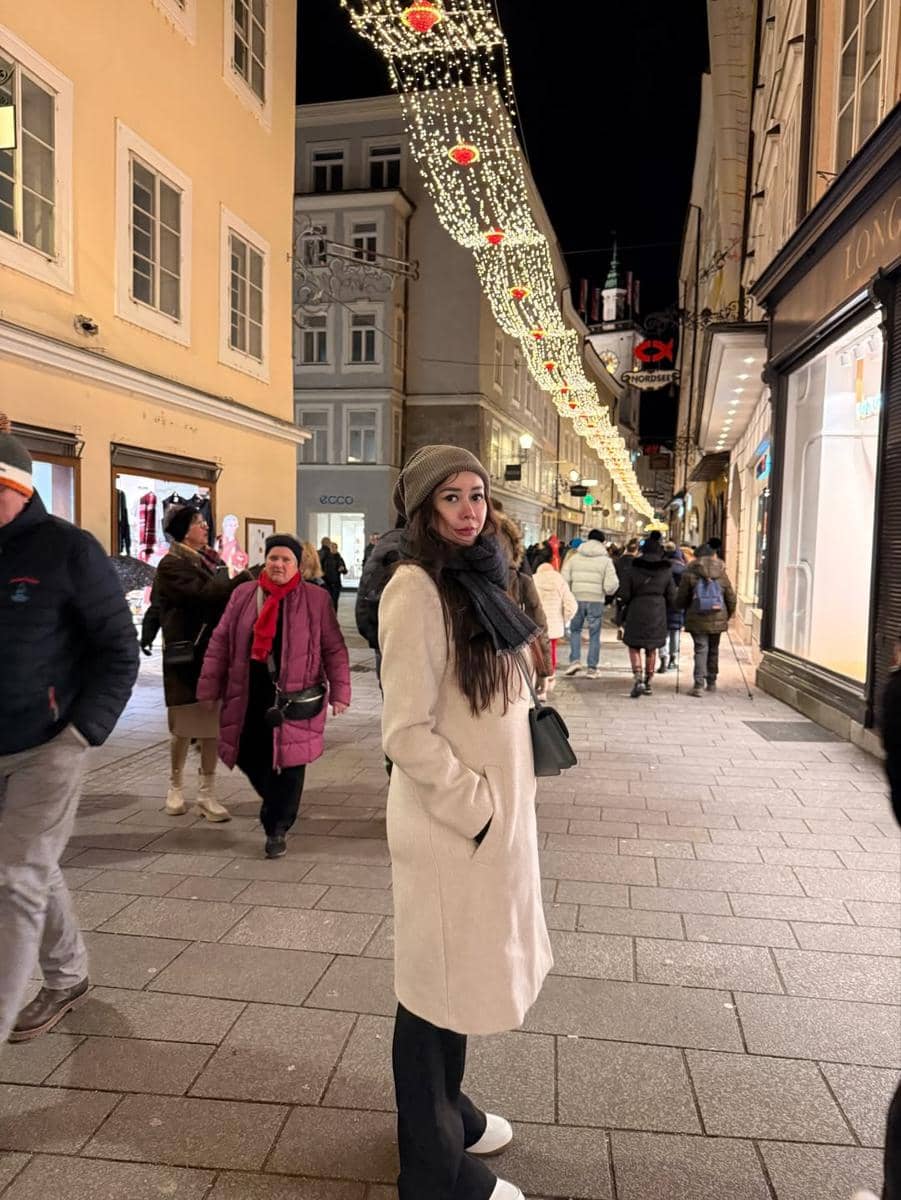 Seorang wanita mengenakan mantel krem dan syal hitam berdiri di jalan kota Austria pada malam hari dengan lampu hias menggantung di atasnya.