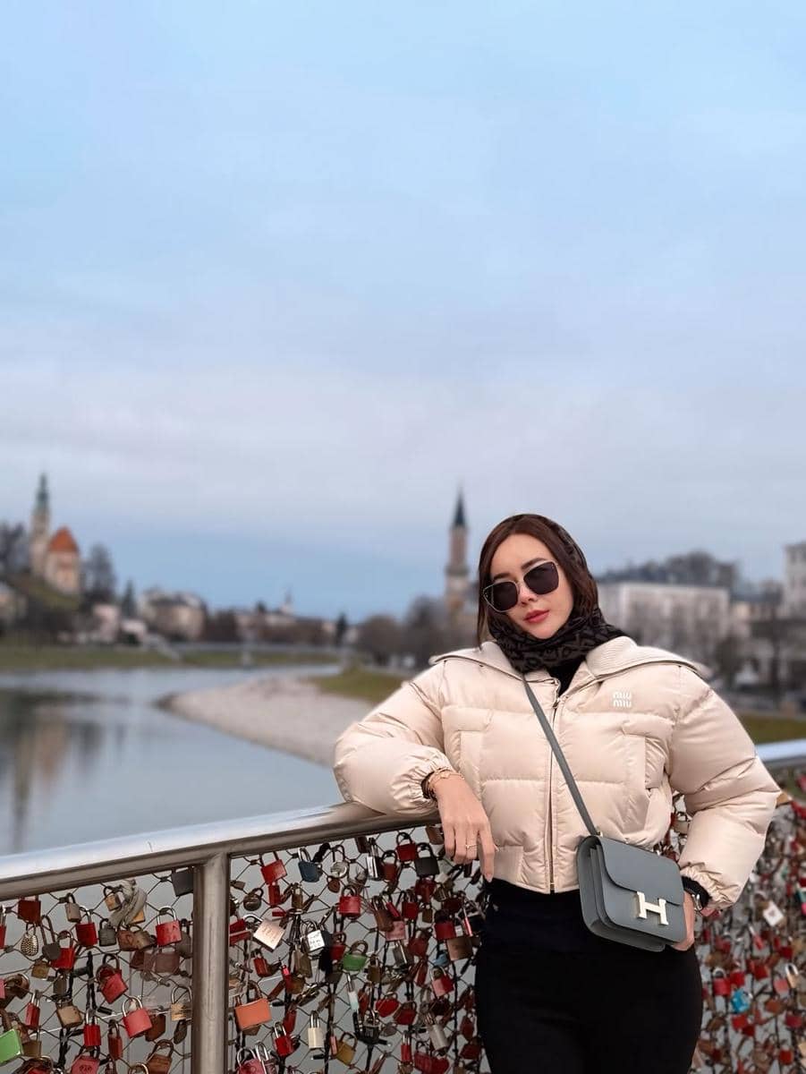 Seorang wanita mengenakan jaket tebal dan kacamata hitam berpose di jembatan penuh gembok cinta dengan latar kota di Austria.