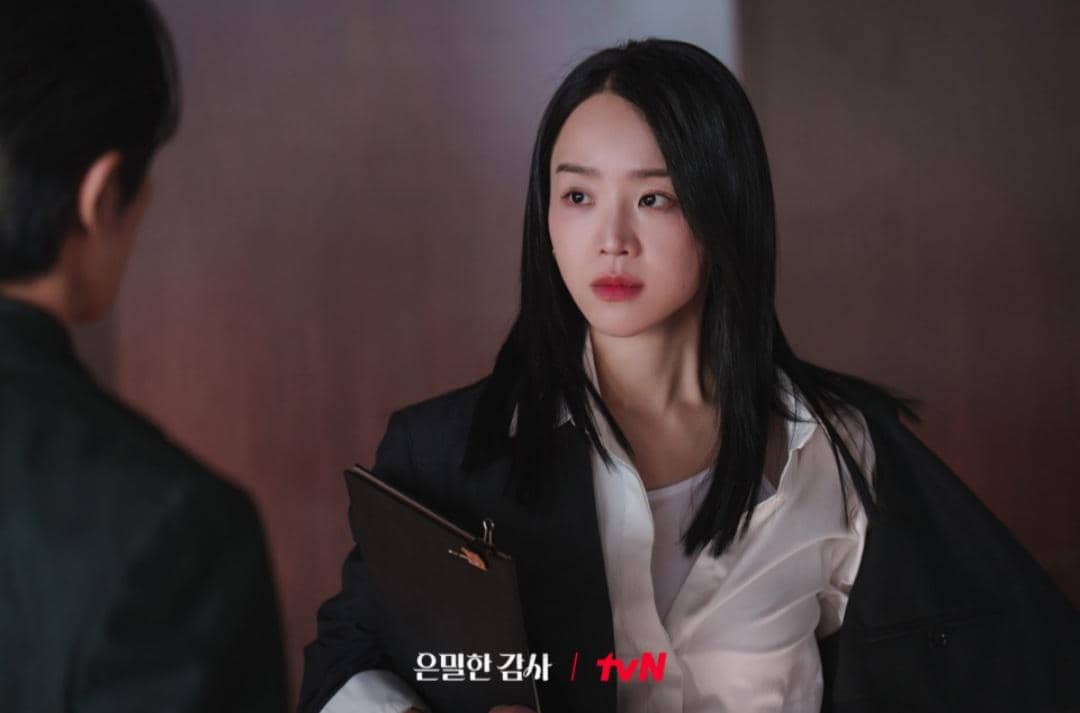 Seorang wanita berambut hitam mengenakan kemeja putih dan jas hitam memegang map, menatap seseorang di depannya dalam adegan drama Korea Filing for Love.