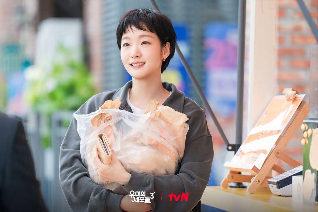 Seorang wanita berambut pendek tersenyum sambil memeluk kantong belanja besar di adegan drama Yumi’s Cells 3.
