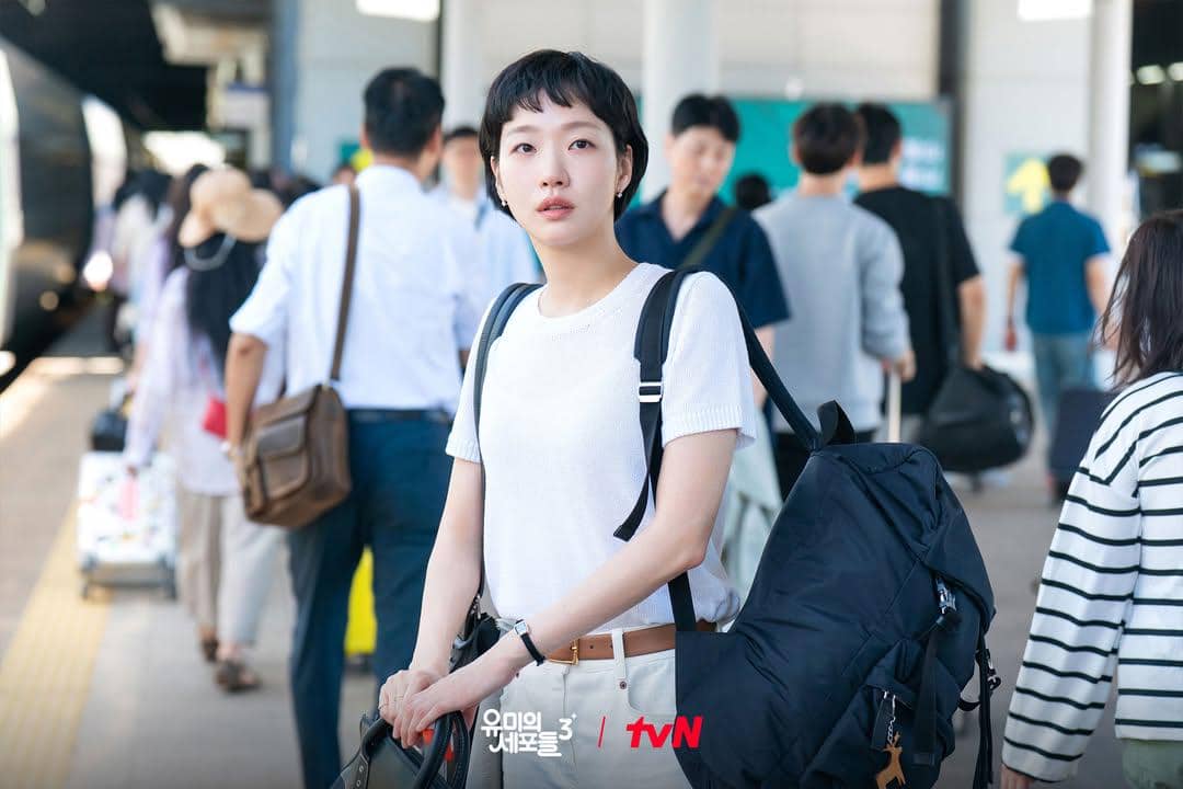 Seorang wanita berambut pendek mengenakan kaus putih dan membawa tas besar berdiri di peron stasiun yang ramai penumpang.
