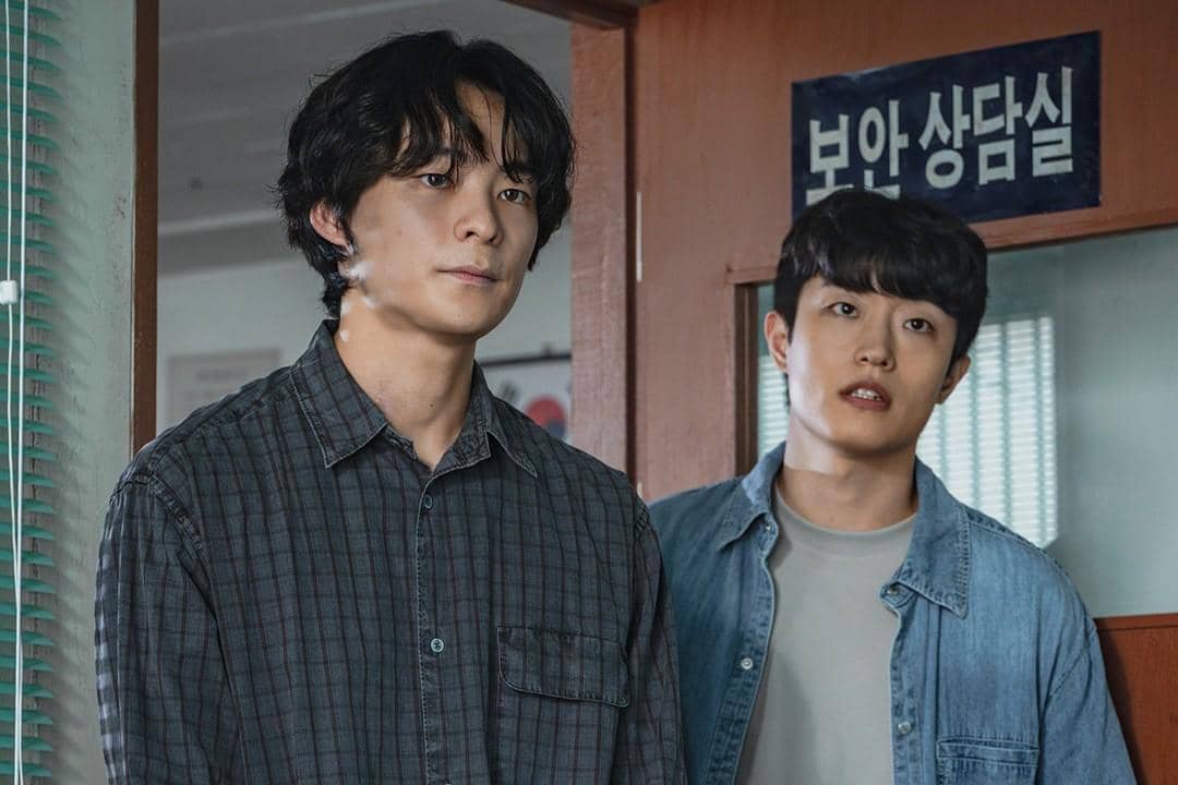 Dua pria berdiri di depan pintu bertuliskan huruf Korea dalam adegan drama The Scarecrow, mengenakan kemeja kasual berwarna gelap dan denim.