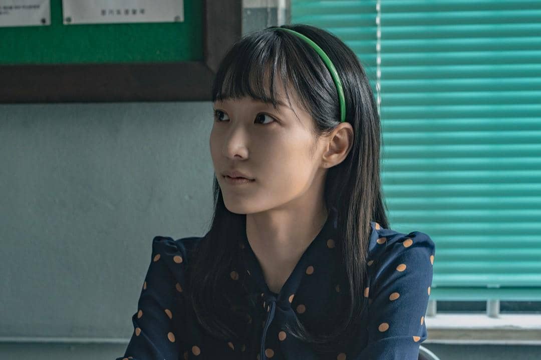 Seorang perempuan berambut panjang mengenakan bando hijau dan blus biru bermotif polkadot duduk di dalam ruangan dengan tirai hijau.