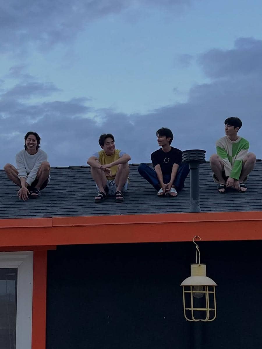 Empat pria duduk di atap rumah dengan langit senja di latar belakang, tampak santai dan tersenyum satu sama lain.