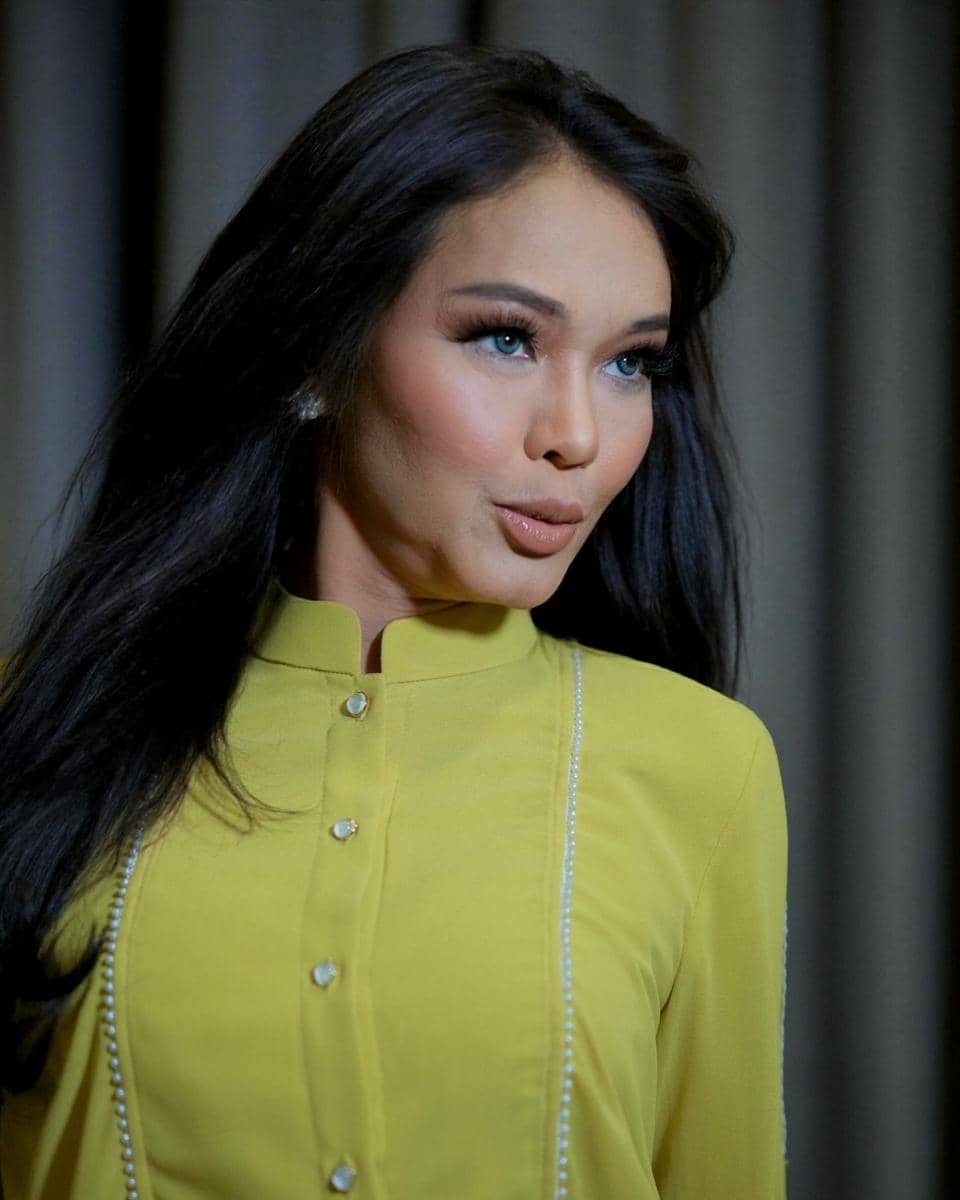 Seorang perempuan berambut panjang mengenakan busana kuning dengan kancing depan, berdiri di depan latar tirai abu-abu.