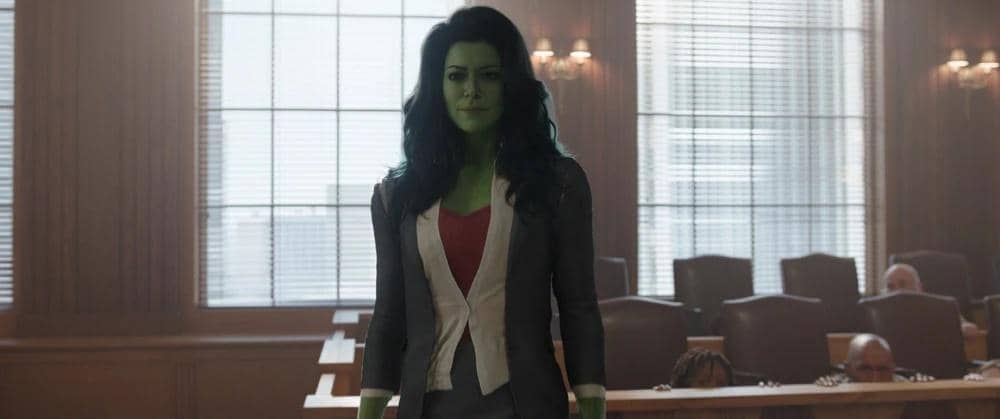 Karakter She-Hulk berdiri di ruang sidang dengan mengenakan setelan jas abu-abu dan blus merah, cahaya masuk melalui jendela besar di belakangnya.