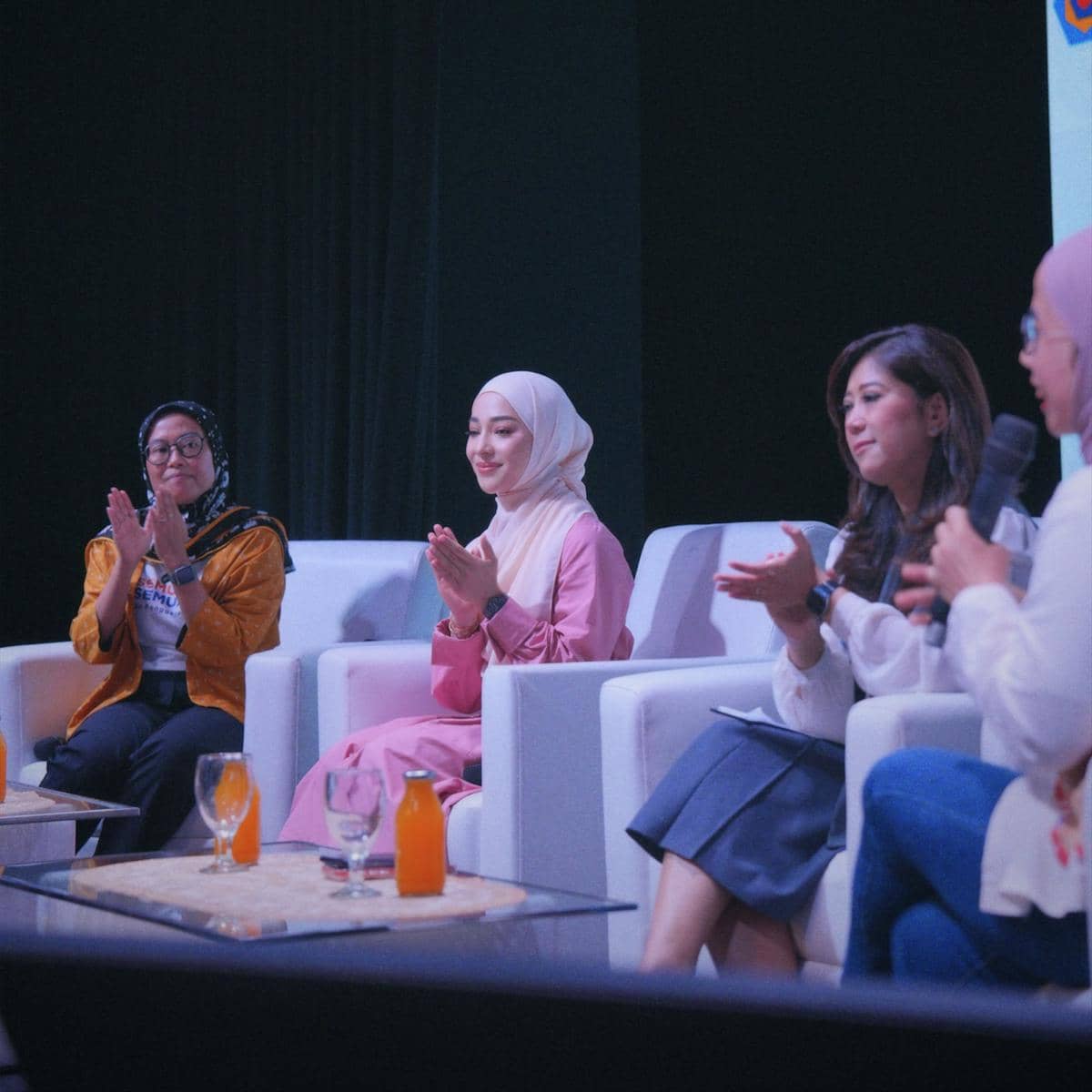 Empat perempuan duduk di kursi putih dalam sebuah diskusi panel, mengenakan pakaian formal dan bertepuk tangan di atas panggung.