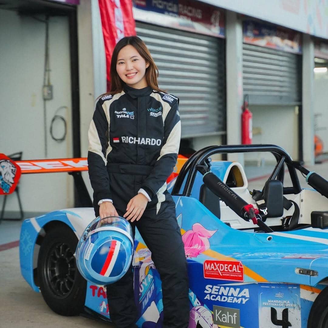 Seorang pembalap perempuan mengenakan baju balap hitam dengan nama Richard di dada, berdiri di samping mobil balap berwarna biru dengan desain animasi.