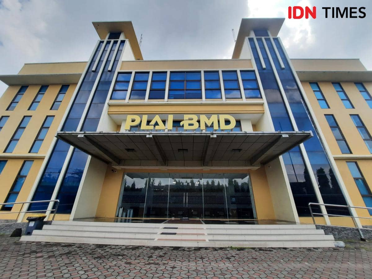 Tampak depan gedung kampus Politeknik AI Budi Mulia Dua dengan arsitektur modern berwarna kuning dan biru serta tulisan PLAI BMD di fasadnya.