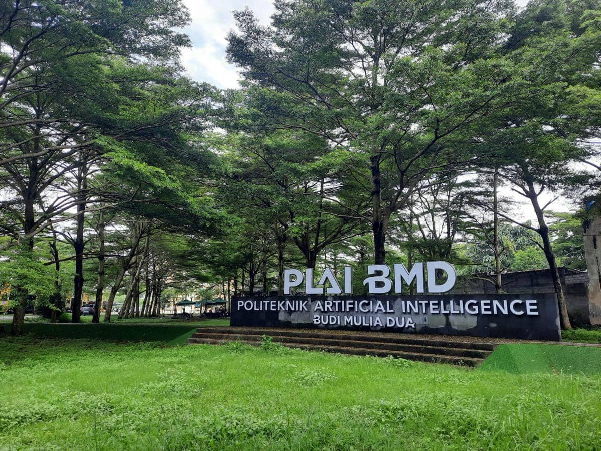 Tampak area hijau dengan pepohonan rindang di depan papan nama Politeknik Artificial Intelligence Budi Mulia Dua (PLAI BMD).