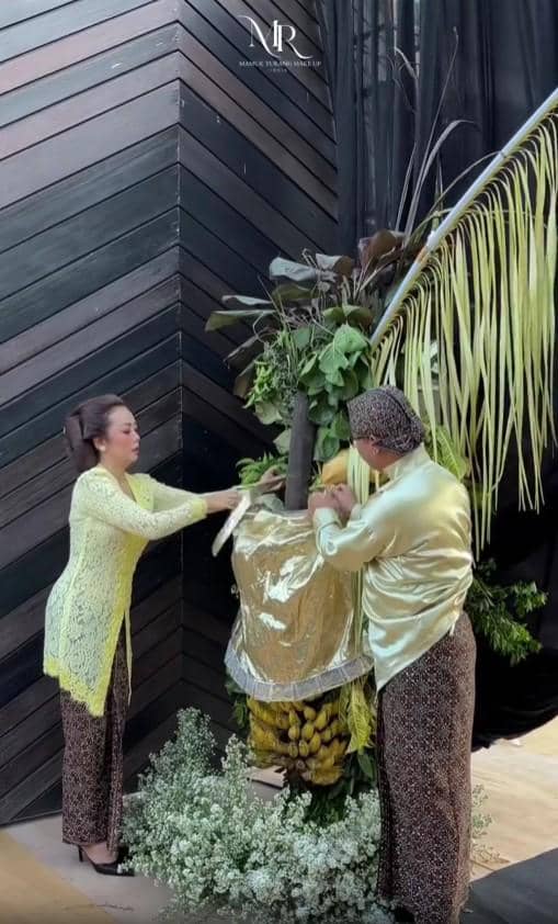 Dua orang mengenakan busana tradisional Jawa sedang menata pohon pisang berhias daun dan bunga dalam prosesi adat menjelang pernikahan.