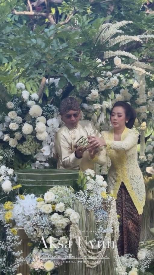 Seorang pria dan wanita mengenakan busana tradisional Jawa berwarna krem dan kuning dalam prosesi siraman di tengah dekorasi bunga putih dan hijau.