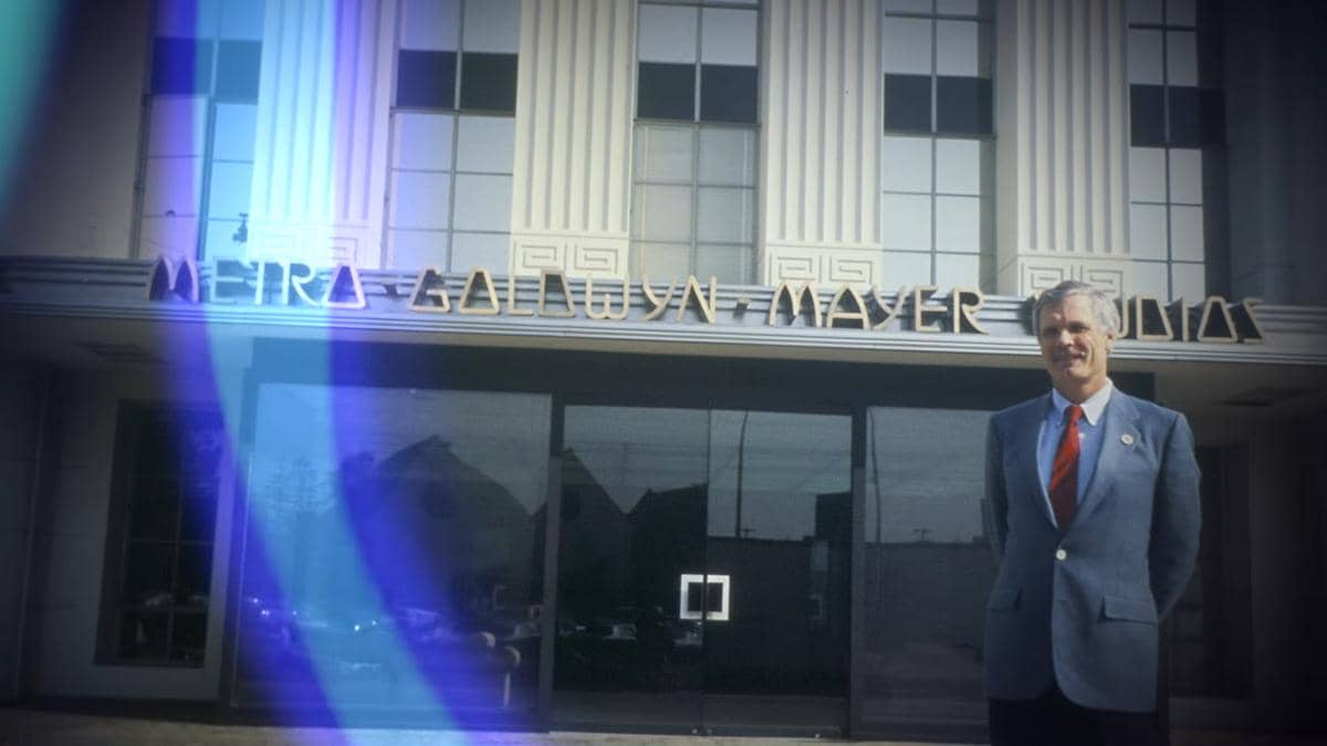 Seorang pria berjas biru dan dasi merah berdiri di depan gedung Metro-Goldwyn-Mayer Studios dengan arsitektur klasik.