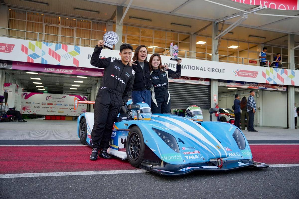 Tiga orang pembalap berpose dengan piala di depan mobil balap biru di sirkuit Mandalika setelah kompetisi motorsport.