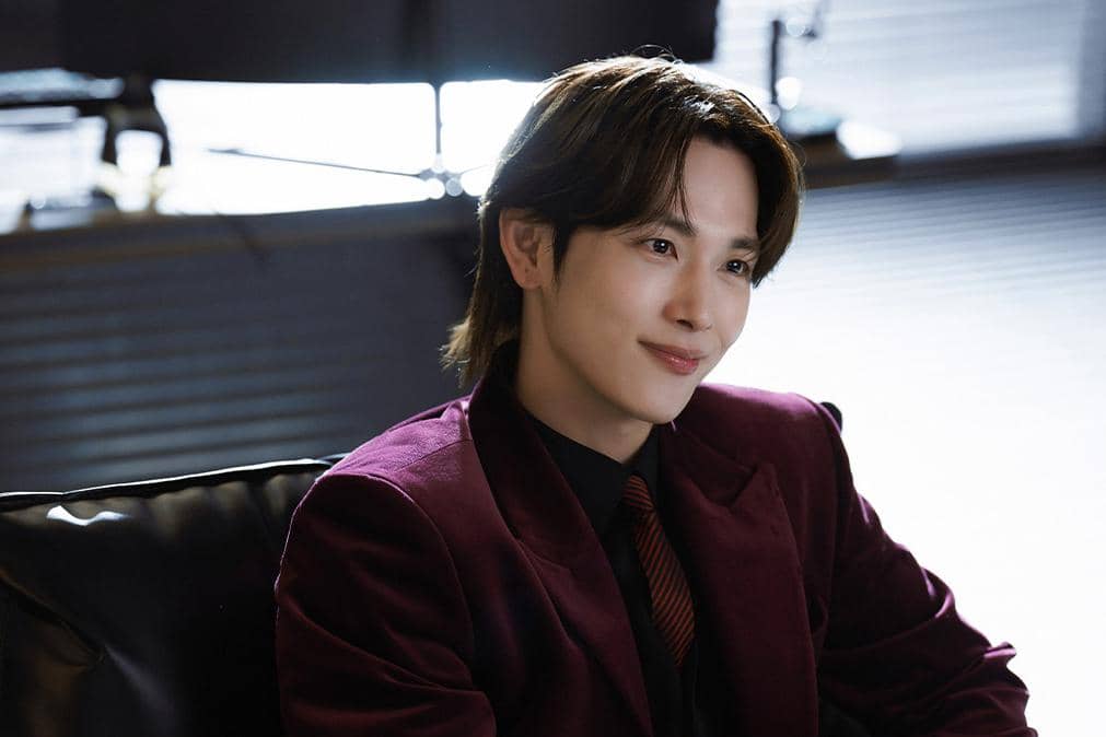 Seorang aktor mengenakan setelan jas merah marun dan dasi hitam-merah duduk tersenyum di kursi dalam adegan drama Korea Love in Disguise.