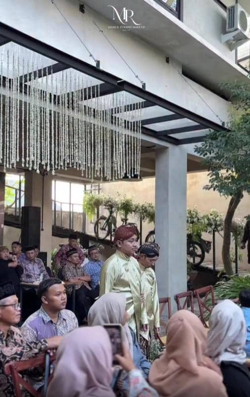 Seorang pria mengenakan pakaian adat Jawa berwarna hijau muda berjalan di tengah acara siraman dan sungkeman menjelang pernikahan.