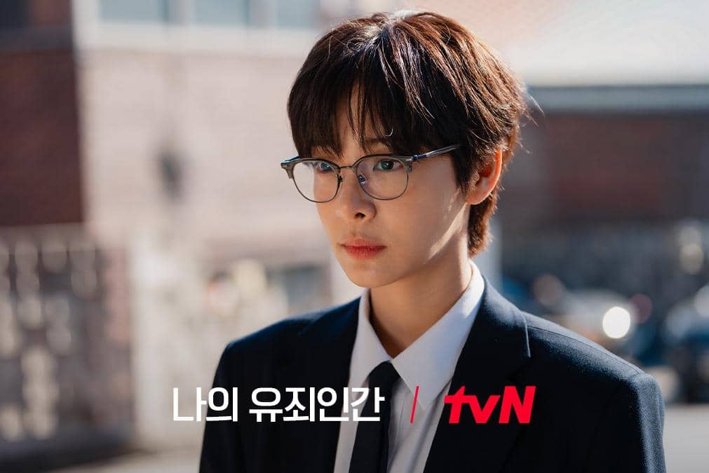 Seorang karakter mengenakan setelan jas hitam dan kacamata berdiri di luar ruangan dengan latar bangunan bata dalam drama Korea Love in Disguise.