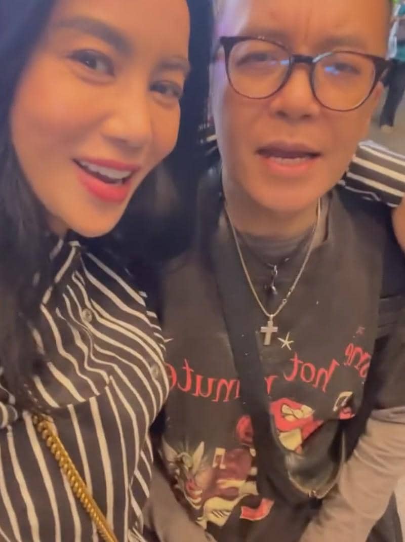 Dua orang berpose bersama sambil tersenyum, salah satunya mengenakan kemeja bergaris hitam putih dan yang lain memakai kacamata serta kalung salib.