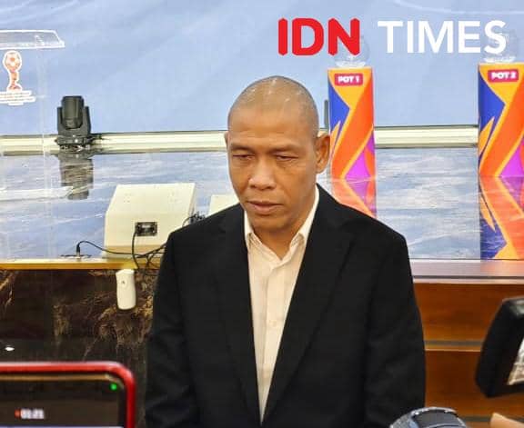 Seorang pria berkemeja putih dan mengenakan jas hitam duduk di ruangan dengan latar belakang meja dan perlengkapan elektronik.