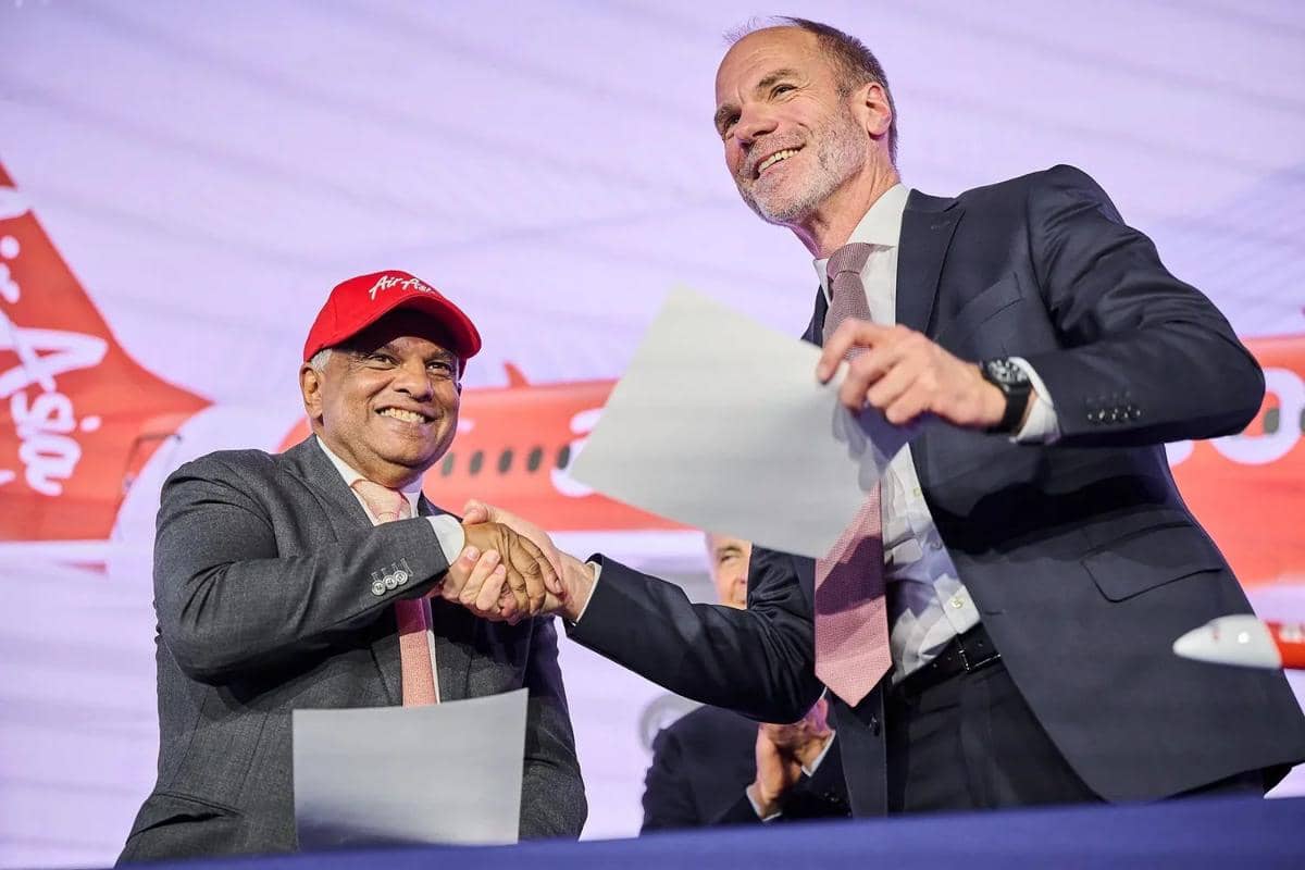 Dua eksekutif berjabat tangan sambil memegang dokumen di depan latar belakang pesawat AirAsia, menandai kesepakatan bisnis besar.