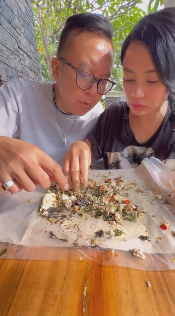 Seorang pria dan wanita duduk berdampingan di meja kayu, menikmati makanan tradisional yang disajikan di atas kertas plastik.