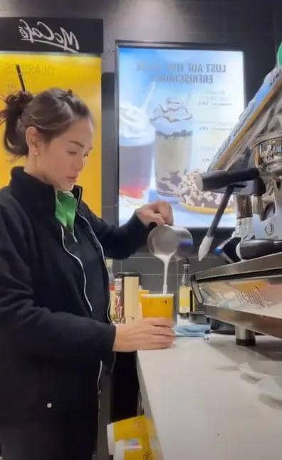 Seorang barista menuangkan susu ke dalam gelas minuman di meja bar kopi dengan mesin espresso di latar belakang.