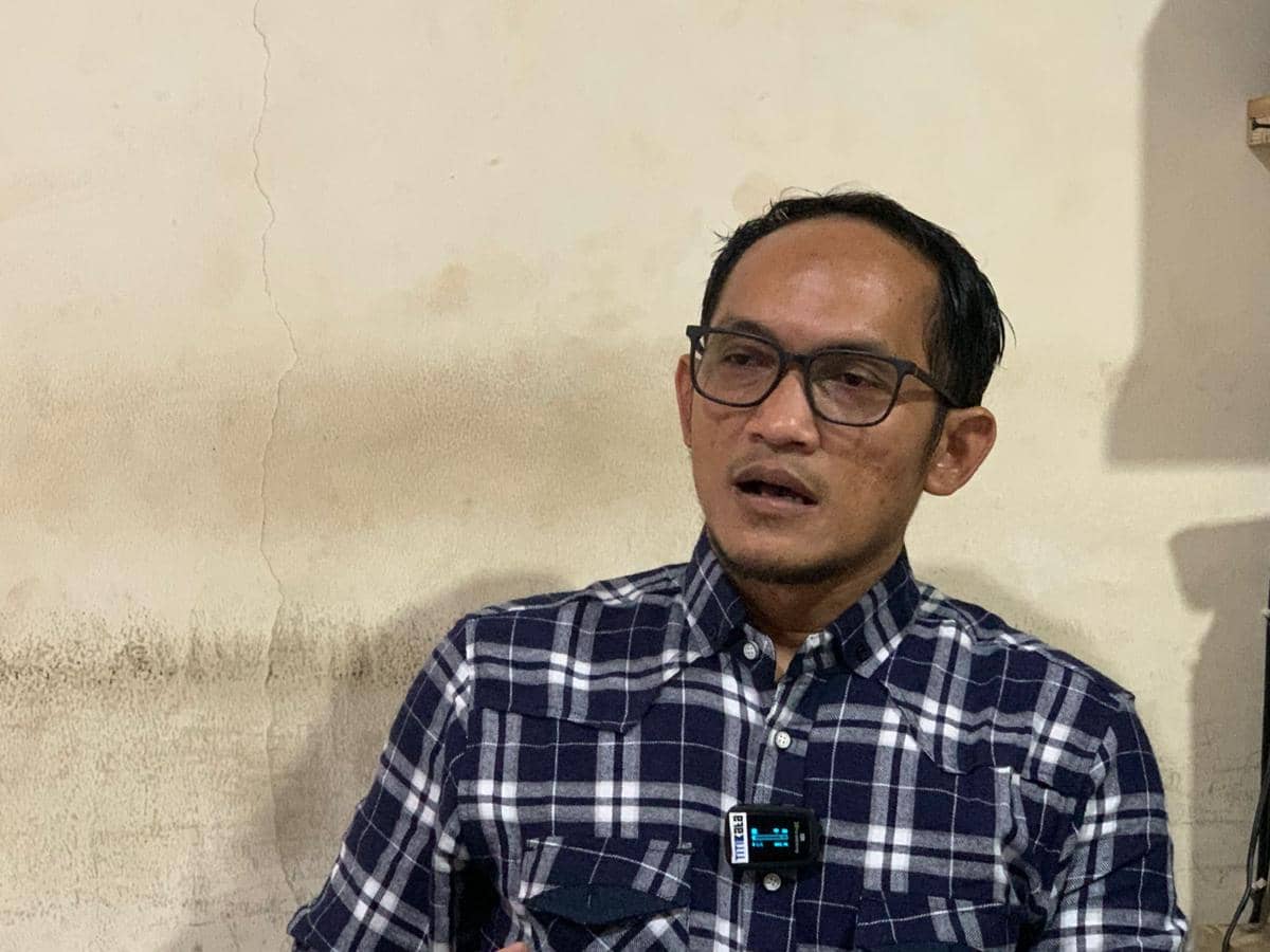 Seorang pria berkacamata mengenakan kemeja kotak-kotak biru duduk di dalam ruangan dengan dinding berwarna krem.