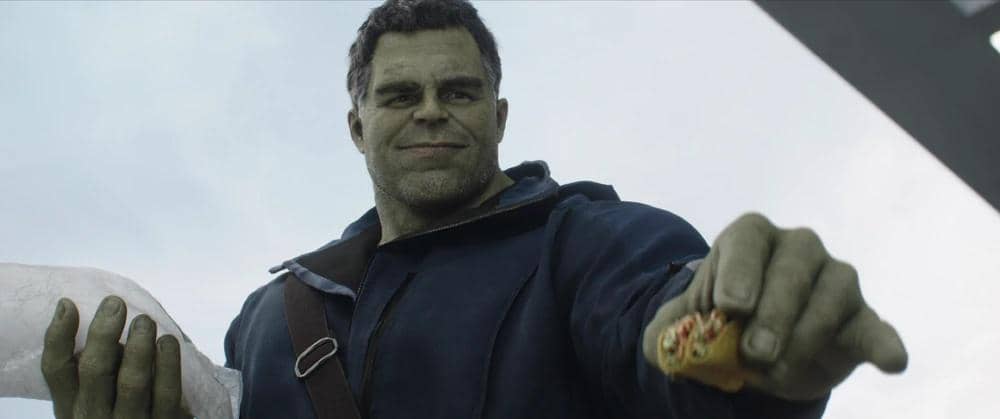 Hulk mengenakan jaket biru sambil memegang makanan di tangan kanan dan roti besar di tangan kiri dengan latar langit cerah.