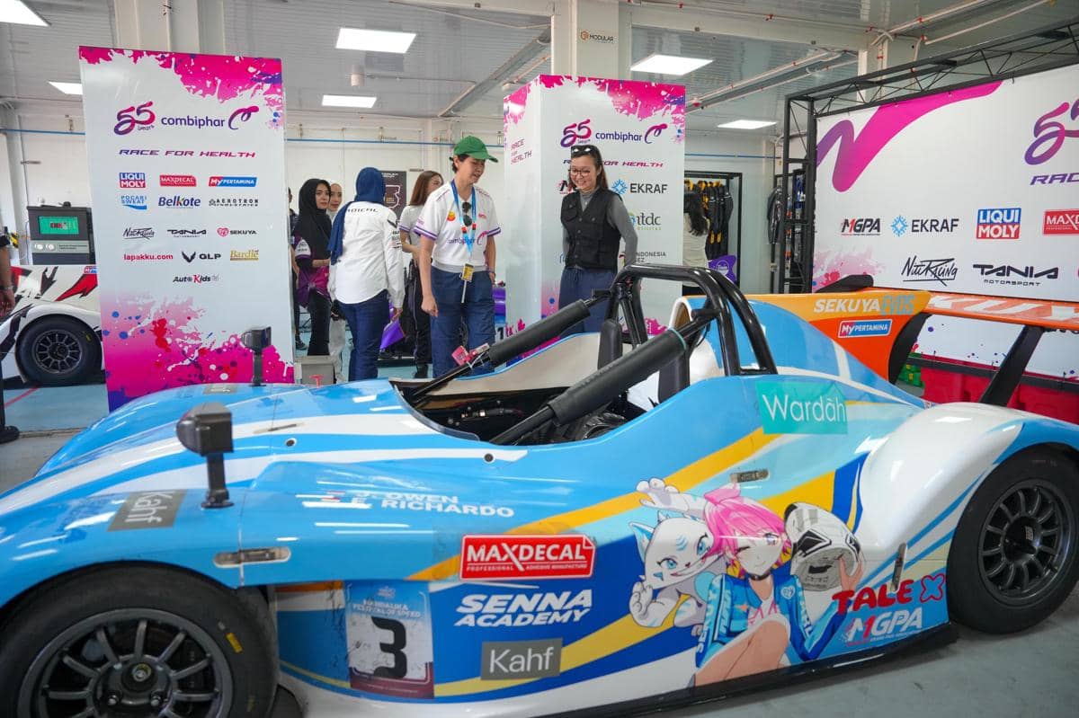 Mobil balap berwarna biru dengan livery Maxdecal bergambar karakter animasi dipamerkan di area pit bersama beberapa orang pengunjung.