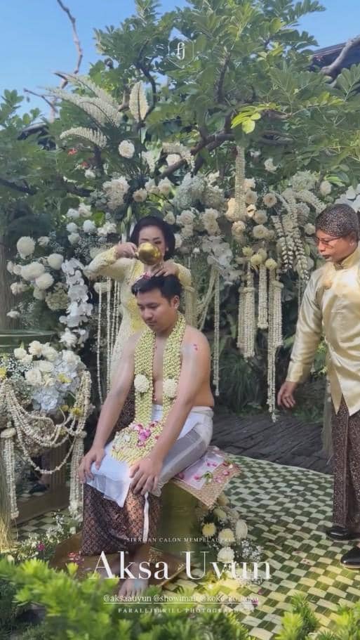 Seorang pria menjalani prosesi siraman adat Jawa dengan air yang dituangkan oleh seorang wanita di taman berhias bunga putih.