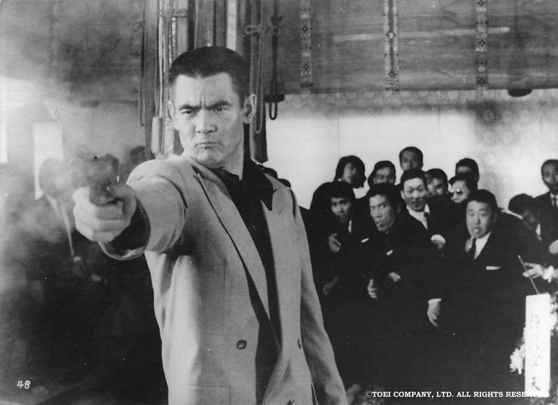 Adegan hitam putih dari film Battles Without Honor and Humanity menampilkan seorang pria menodongkan pistol di ruangan penuh orang.
